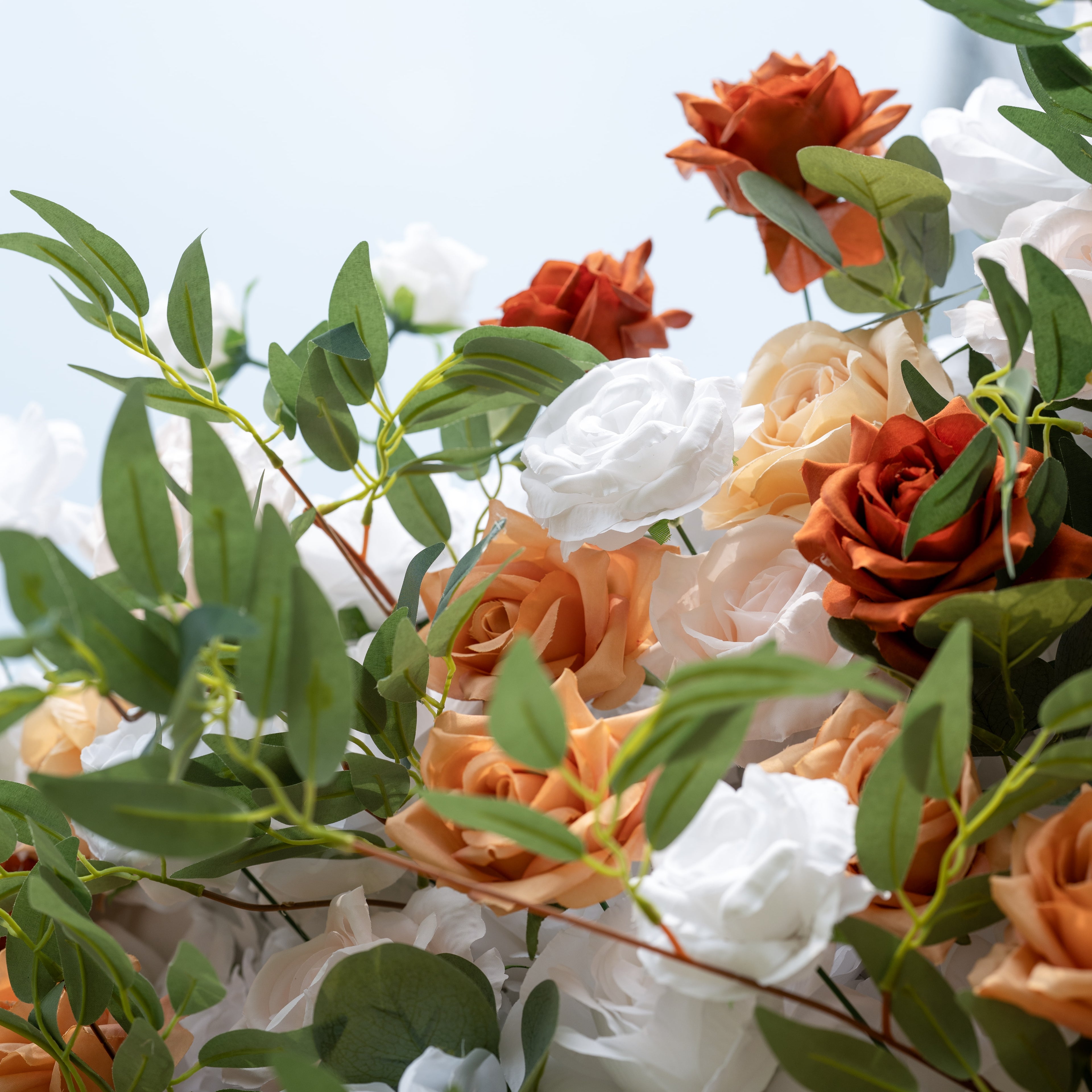 This is a close-up of a premium artificial flower arrangement, brimming with lifelike blooms in a warm terracotta and cream palette. It features burnt orange roses, creamy white roses, blush peach roses, and realistic eucalyptus greenery. The high-quality, realistic petals and foliage create a fresh, timeless, romantic aesthetic, perfect for wedding backdrops, photo booths, fall events, and luxury event decor.