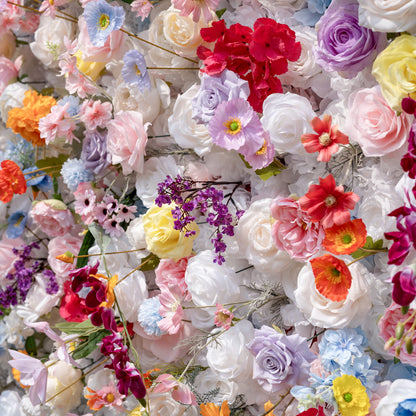This is a lush, vibrant close-up of a premium artificial mixed-flower wall, brimming with lifelike blooms in a joyful rainbow palette. It features creamy white roses, soft pink peonies, sunny yellow roses, vivid orange carnations, sky-blue poppies, lavender roses, deep red hydrangeas, and delicate purple filler flowers. The layered, realistic petals create a dreamy, romantic, whimsical aesthetic, perfect for wedding backdrops, event decor, and photo booth styling.