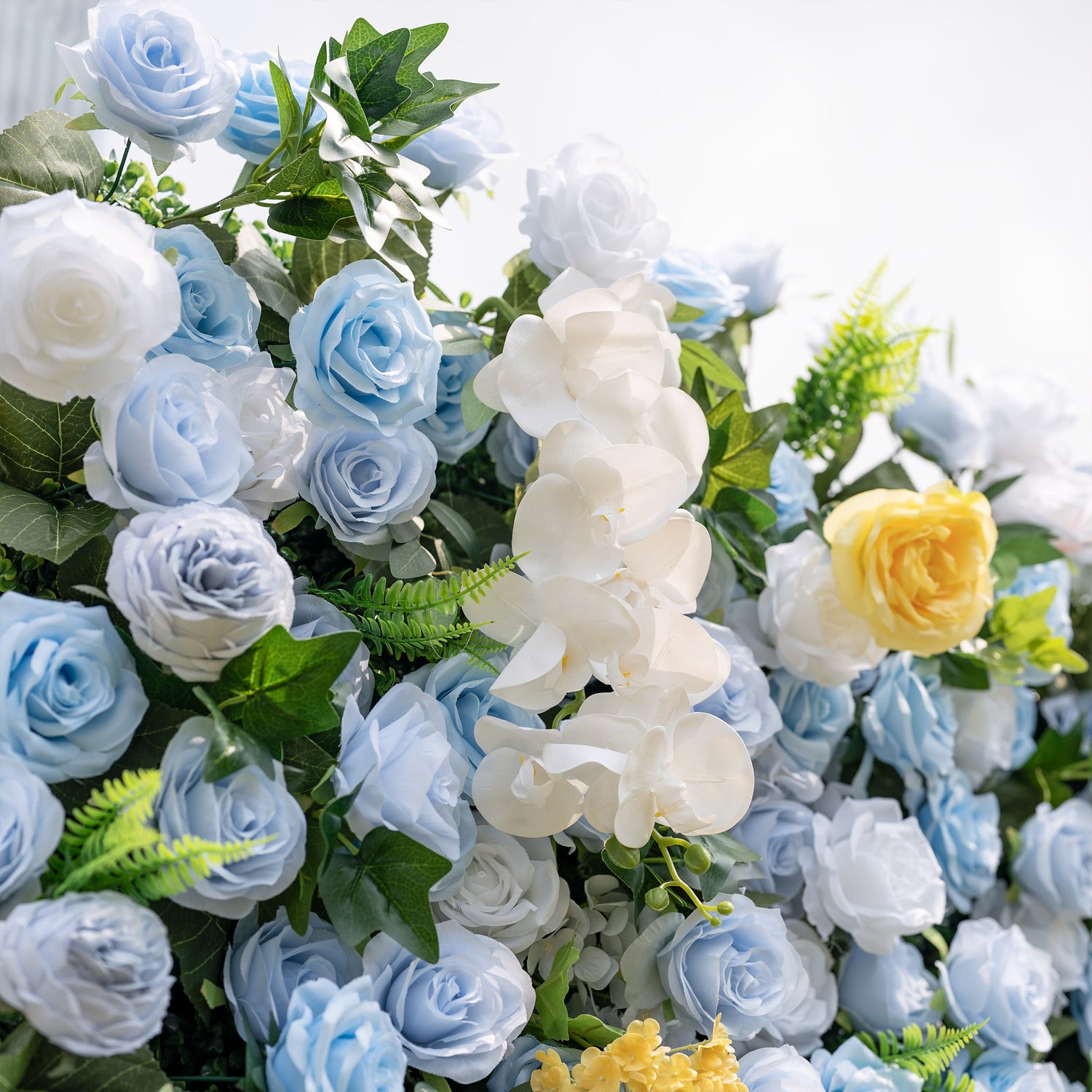 This is a lush, premium artificial flower wall arrangement, brimming with lifelike blooms in a dreamy cool-toned palette. It features sky-blue roses, creamy white roses, elegant white phalaenopsis orchids, sunny yellow roses, and vibrant green foliage. The dense, realistic design creates a fresh, elegant, romantic focal point, perfect for wedding backdrops, photo booths, bridal showers, and luxury event decor.