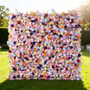 This image features a large, square floral wall outdoors on a lush green lawn. It's densely covered with an array of vibrant flowers in a wide spectrum of colors, including pink, purple, blue, orange, and yellow, creating a lively and eye-catching display. Bathed in sunlight, with trees and a clear sky in the background, the scene exudes a sense of natural beauty and festive charm, perfect for events or as a stunning decorative backdrop.