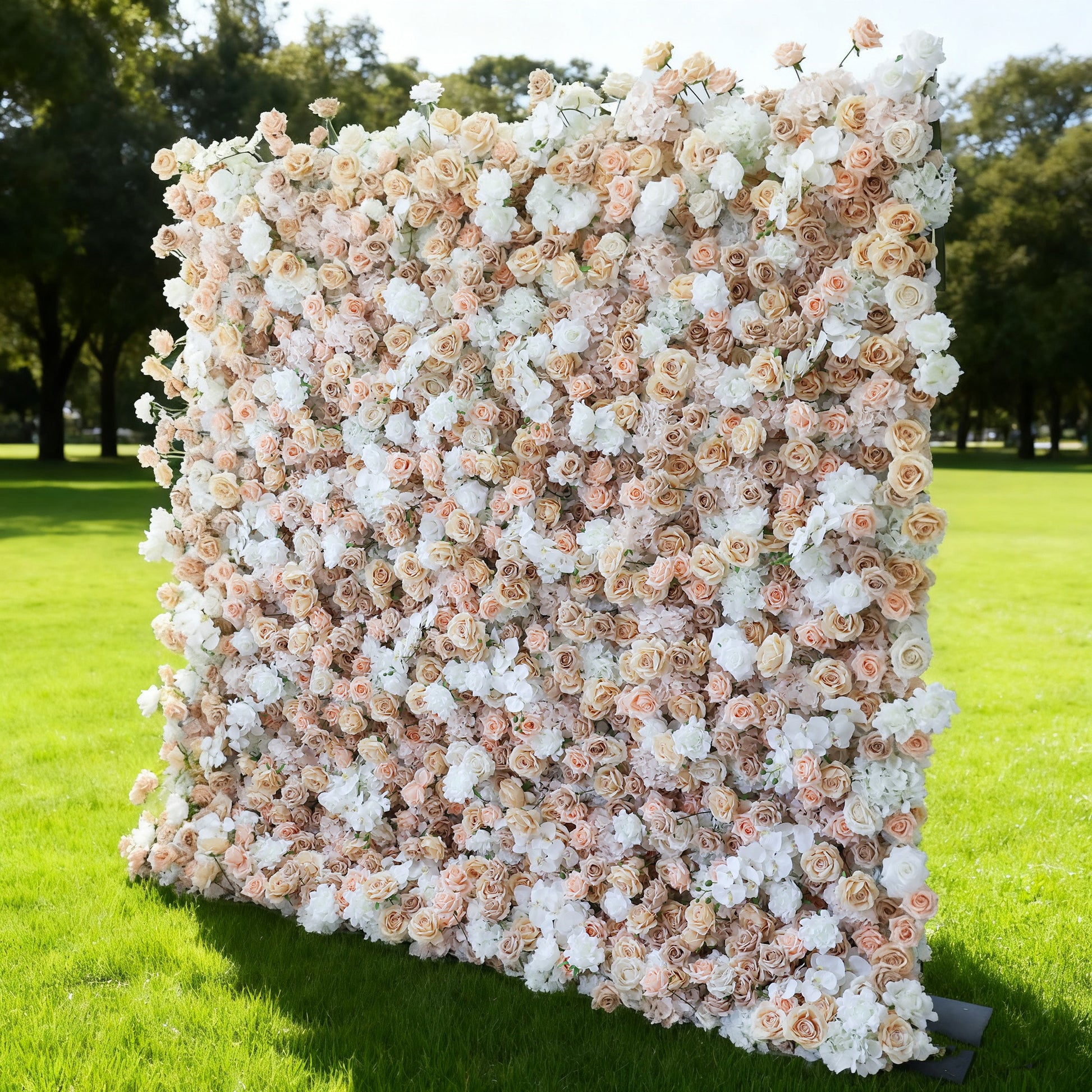 This image features a large, square floral wall outdoors on a lush green lawn. It's densely covered with an array of flowers in soft hues of beige, white, and light pink, creating a romantic and elegant display. Bathed in sunlight, with trees and a clear sky in the background, the scene exudes a sense of natural beauty and charm, perfect for events like weddings or celebrations.