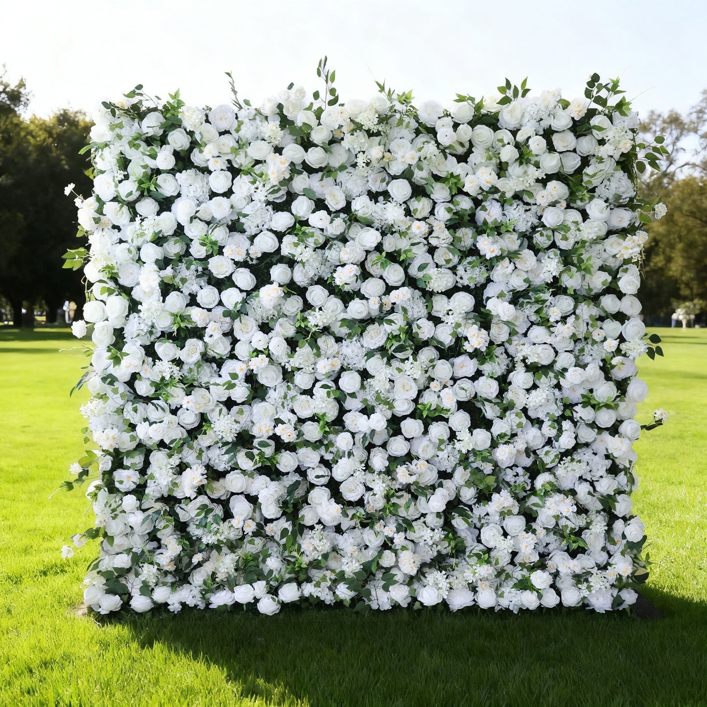 This image features a large, square floral wall outdoors on a lush green lawn. It's densely covered with an array of white flowers, creating a pure and elegant display. Bathed in sunlight, with trees and a clear sky in the background, the scene exudes a sense of natural beauty, purity, and charm, perfect for decorative purposes in events or as a picturesque backdrop.