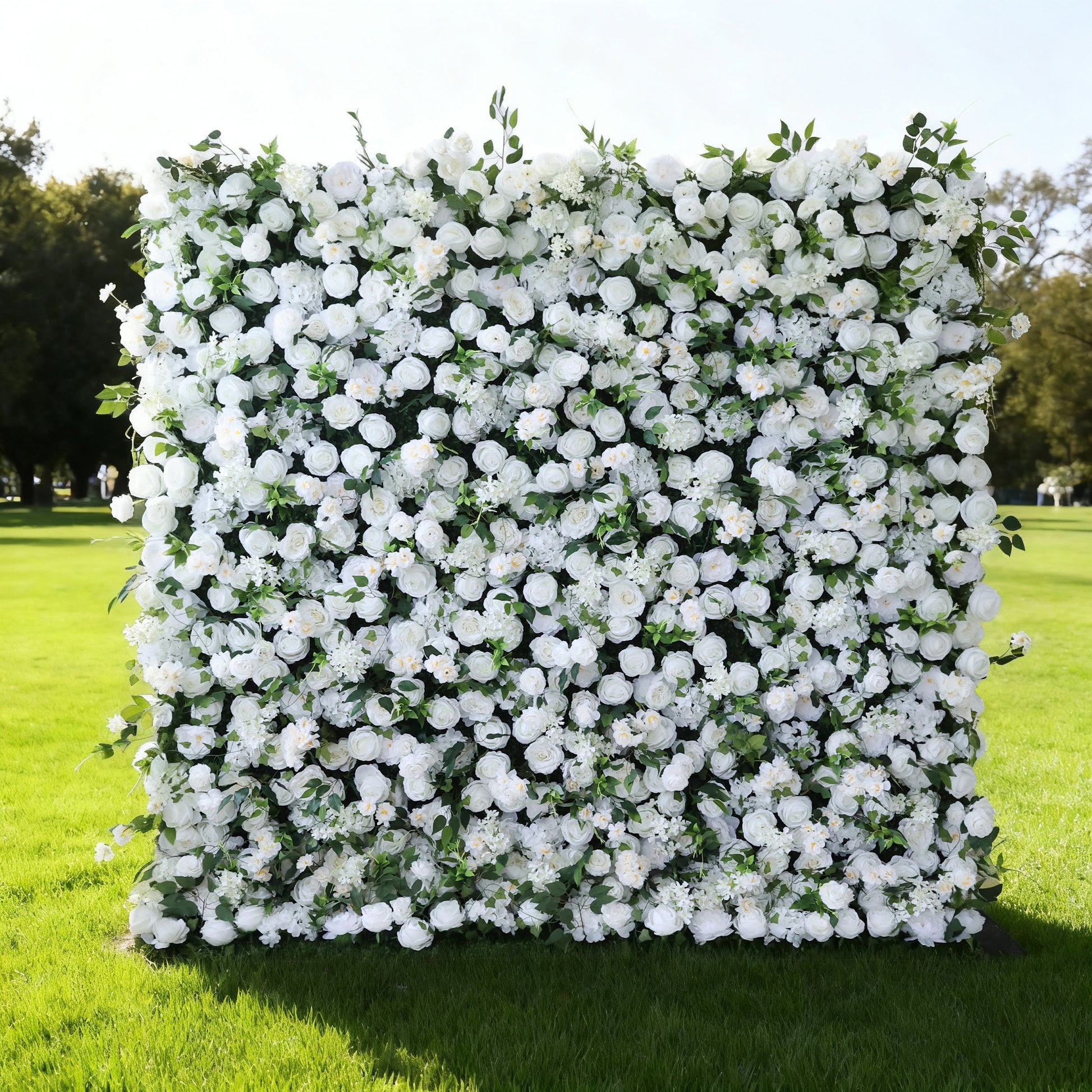 This image features a large, square floral wall outdoors on a lush green lawn. It's densely covered with an array of white flowers, creating a pure and elegant display. Bathed in sunlight, with trees and a clear sky in the background, the scene exudes a sense of natural beauty, purity, and charm, perfect for decorative purposes in events or as a picturesque backdrop.