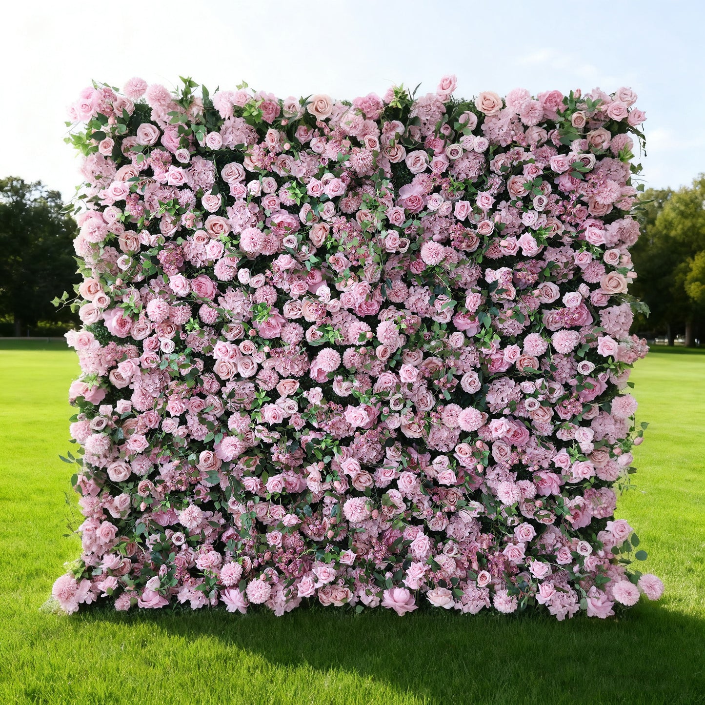 This image features a large, square floral wall outdoors on a lush green lawn. It's densely covered with an array of pink flowers, creating a rich, romantic display. Bathed in sunlight, with trees and a clear sky in the background, the scene exudes a sense of natural beauty, elegance, and charm, perfect for decorative purposes in events or as a picturesque backdrop.