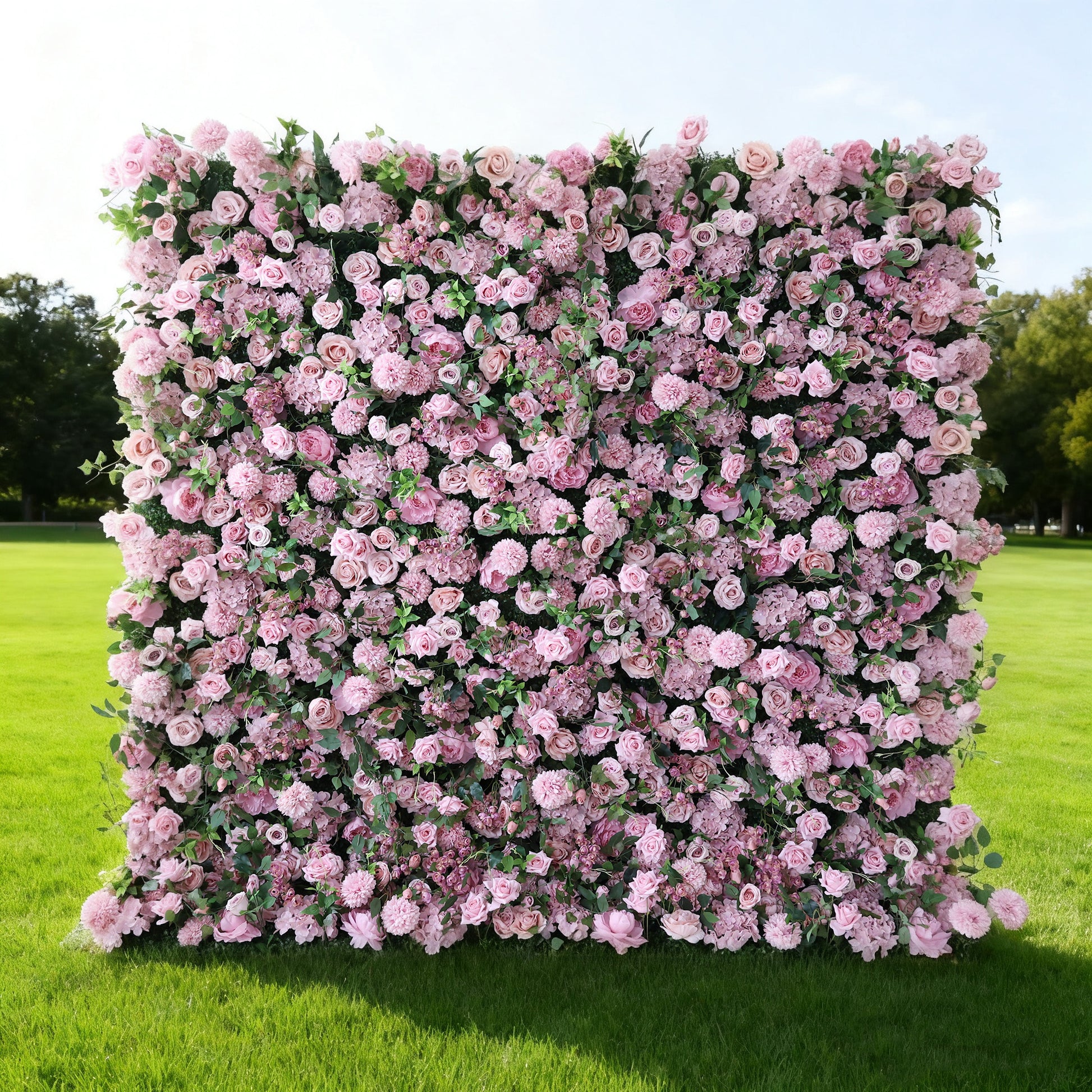 This image features a large, square floral wall outdoors on a lush green lawn. It's densely covered with an array of pink flowers, creating a rich, romantic display. Bathed in sunlight, with trees and a clear sky in the background, the scene exudes a sense of natural beauty, elegance, and charm, perfect for decorative purposes in events or as a picturesque backdrop.