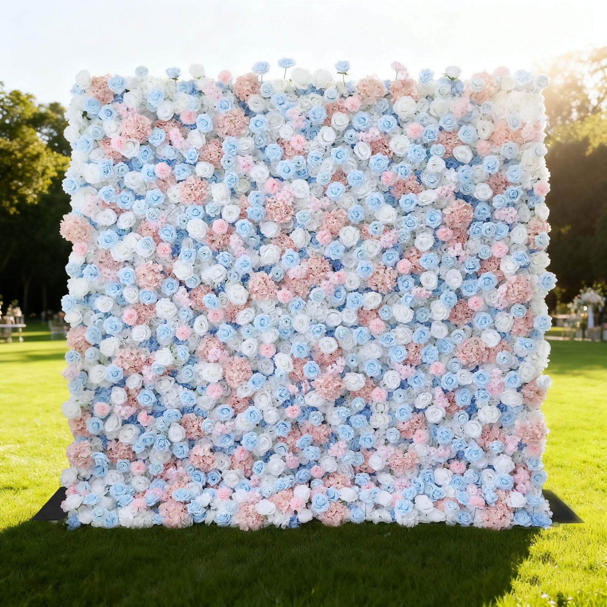This image features a large, square floral wall outdoors on a lush green lawn venue. It's densely covered with an array of flowers in soft pink, light blue, and white hues. The vibrant blooms create a rich, colorful, and romantic display. Bathed in sunlight, with trees and a clear sky in the background, the scene exudes a sense of beauty, elegance, and natural charm, perfect for decorative purposes in events or as a picturesque backdrop.