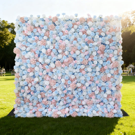This image features a large, square floral wall outdoors on a lush green lawn venue. It's densely covered with an array of flowers in soft pink, light blue, and white hues. The vibrant blooms create a rich, colorful, and romantic display. Bathed in sunlight, with trees and a clear sky in the background, the scene exudes a sense of beauty, elegance, and natural charm, perfect for decorative purposes in events or as a picturesque backdrop.