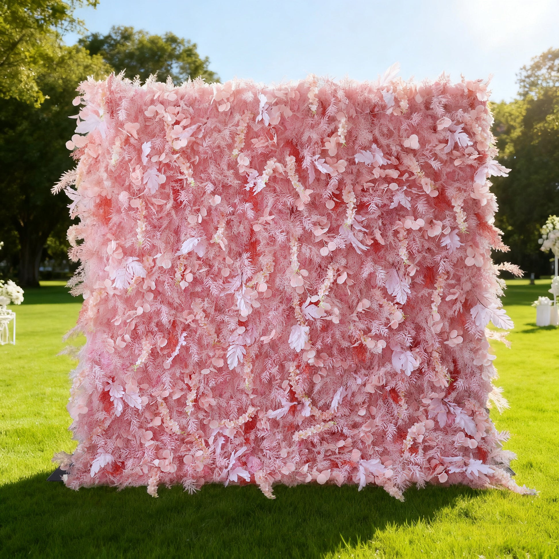 This image features a large, square floral wall outdoors on a lush green lawn. It's densely covered with an array of delicate pink flowers and foliage, creating a soft and romantic display. Bathed in sunlight, with trees and other decorative elements in the background, the scene exudes a sense of natural beauty and gentle charm, perfect for events like weddings or celebrations.
