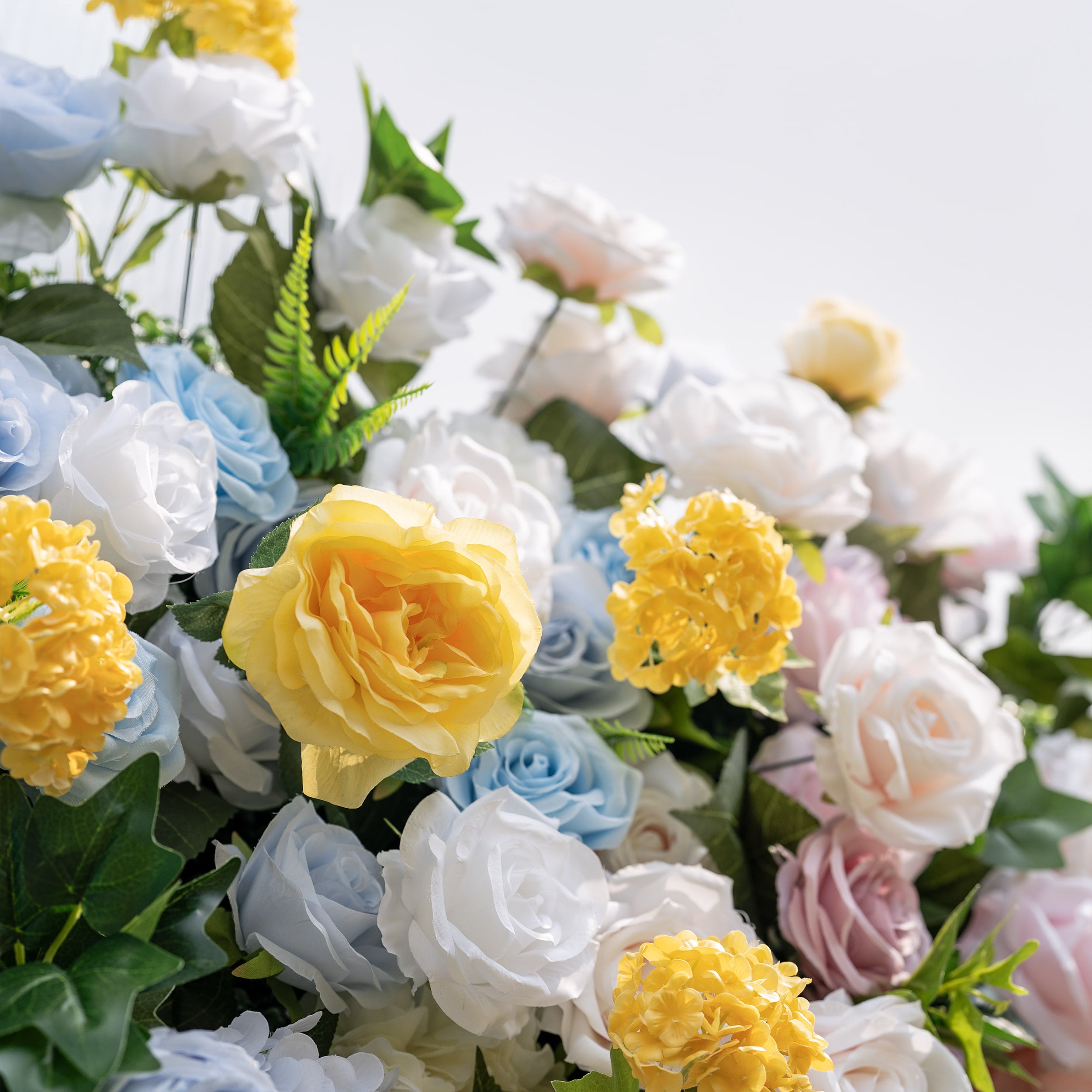 This is a lush, premium artificial flower arrangement, brimming with lifelike blooms in a dreamy pastel palette. It features sky-blue roses, creamy white roses, sunny yellow roses and hydrangeas, blush pink roses, and vibrant green foliage. The dense, realistic design creates a fresh, elegant, romantic focal point, perfect for wedding backdrops, photo booths, bridal showers, and luxury event decor.