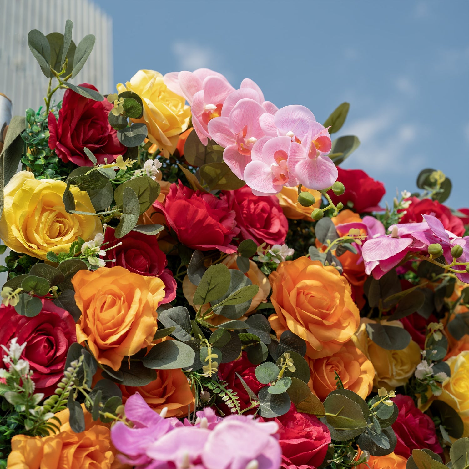 This is a close-up of a premium multicolor artificial flower arrangement, brimming with lifelike blooms: vibrant red, orange, yellow roses, delicate pink phalaenopsis orchids, and realistic eucalyptus greenery with tiny white filler flowers. High-quality, hyper-realistic petals deliver a fresh, lush, long-lasting aesthetic, perfect for wedding backdrops, artificial flower walls, photo booths, summer events, and luxury event decor.