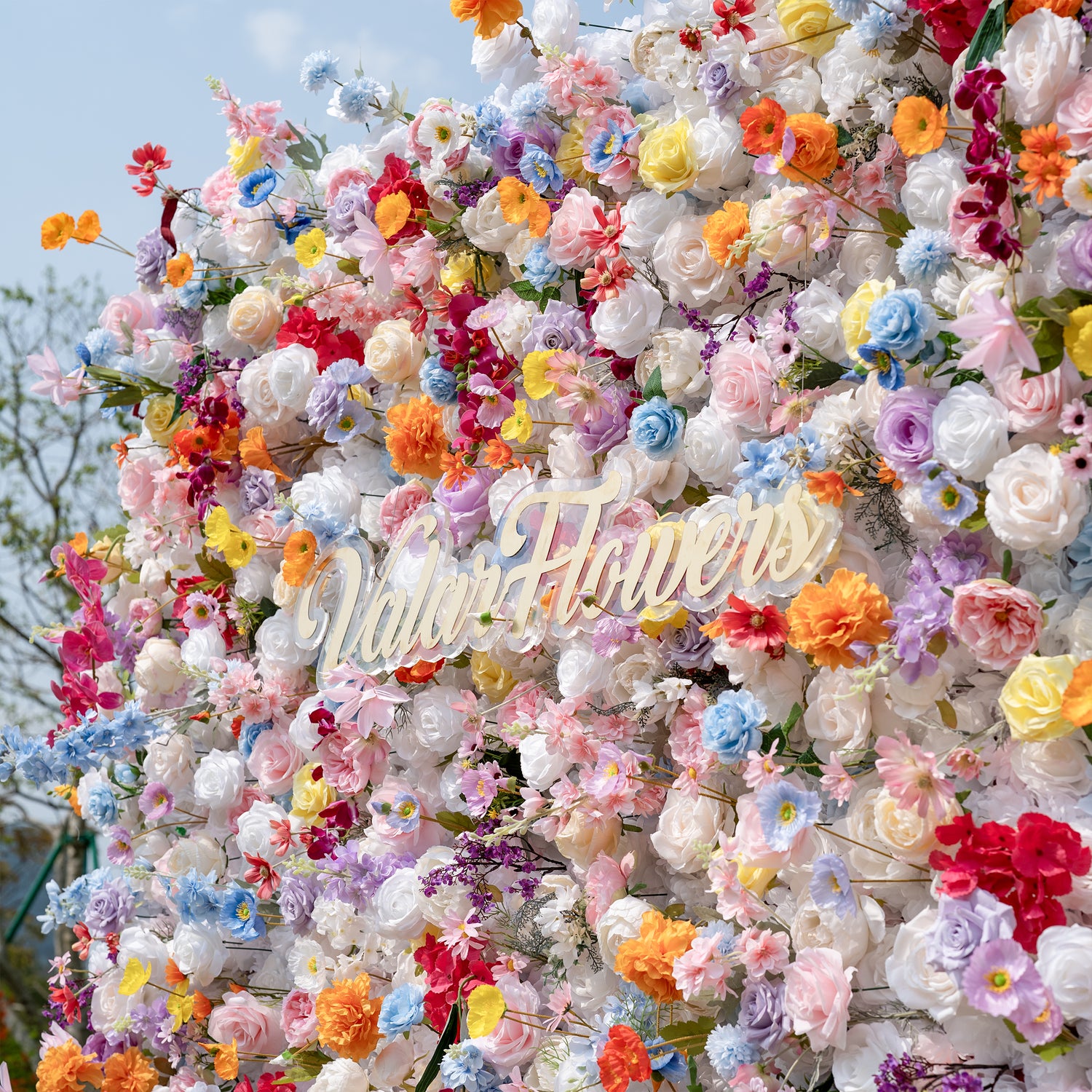 This is a stunning, dense artificial flower wall from Valar Flowers, brimming with a vibrant, rainbow-hued mix of lifelike blooms. It features creamy white roses, soft pink peonies, sunny yellow poppies, vivid orange carnations, sky-blue delphiniums, deep magenta orchids, and delicate filler flowers, set against a bright blue sky. The lush, layered arrangement creates a romantic, showstopping focal point perfect for weddings, photo booths, and luxury event decor.