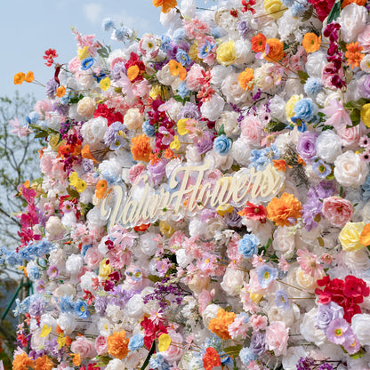 This is a stunning, dense artificial flower wall from Valar Flowers, brimming with a vibrant, rainbow-hued mix of lifelike blooms. It features creamy white roses, soft pink peonies, sunny yellow poppies, vivid orange carnations, sky-blue delphiniums, deep magenta orchids, and delicate filler flowers, set against a bright blue sky. The lush, layered arrangement creates a romantic, showstopping focal point perfect for weddings, photo booths, and luxury event decor.
