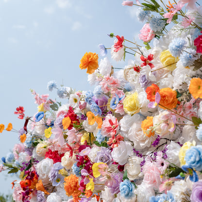 Valar Flowers premium vibrant mixed artificial flower wall backdrop, a lush, layered arrangement of lifelike blooms including white roses, pink peonies, orange carnations, blue poppies, yellow roses, purple orchids, and delicate filler flowers. Set against a bright blue sky, this dense, rainbow-hued floral display creates a dreamy, romantic focal point perfect for outdoor weddings, engagement parties, photo booths, and luxury event decor.