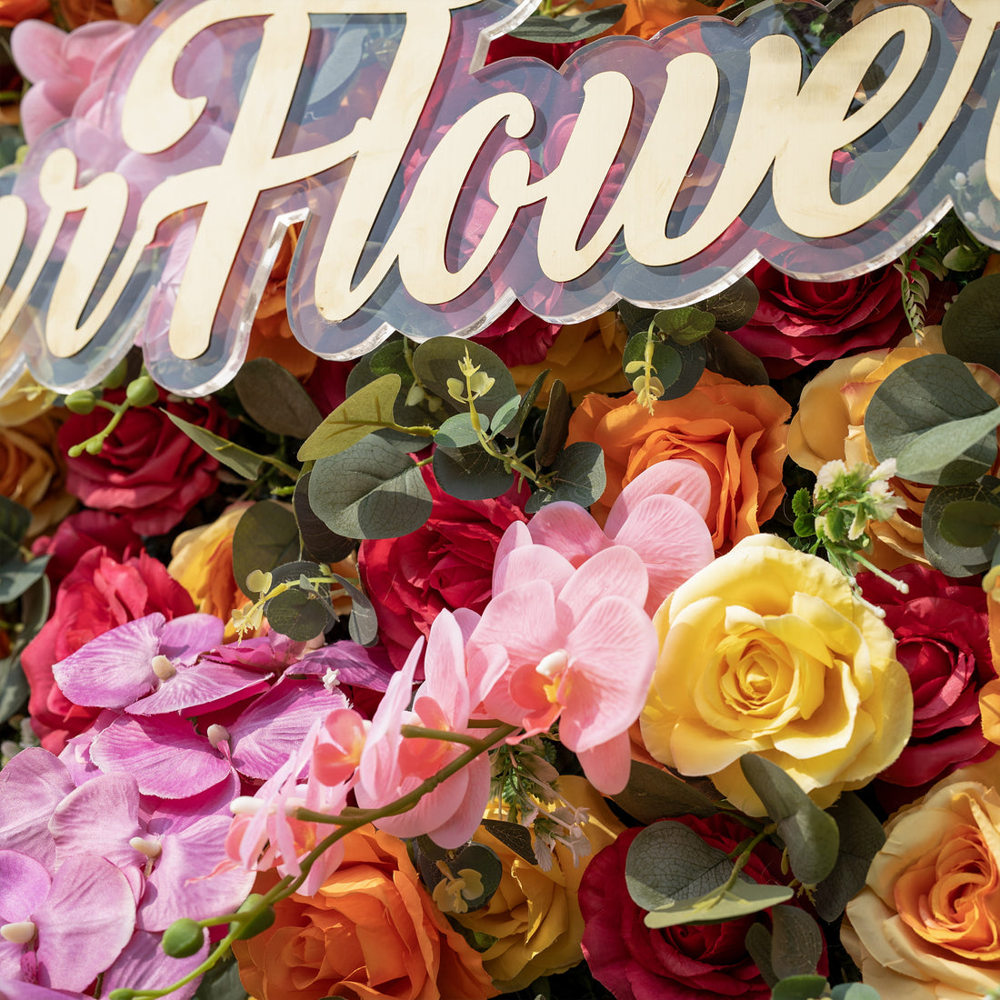 This is a close-up of ValarFlowers’ premium multicolor artificial flower wall, featuring a lush, dense arrangement of lifelike red, orange, yellow roses, pink phalaenopsis orchids, and realistic eucalyptus greenery. Centered is a custom acrylic "ValarFlowers" logo sign. This high-quality, reusable floral backdrop delivers a vibrant, fresh aesthetic, ideal for wedding backdrops, photo booths, summer events, and luxury event decor.