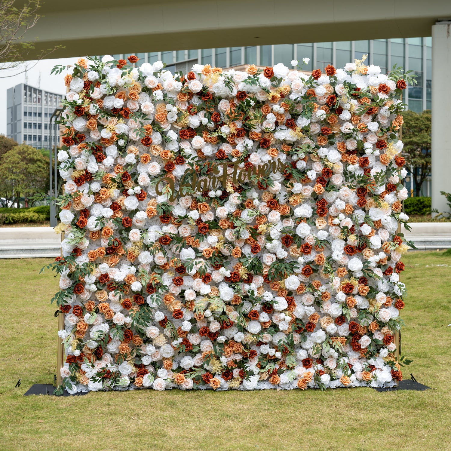 This is a stunning 8x8ft (2.4x2.4m) artificial flower wall backdrop, mounted on a golden metal stand and set outdoors on a grassy lawn with an urban skyline in the background. It features a lush, dense arrangement of lifelike blooms in a warm terracotta and cream palette: burnt orange roses, creamy white peonies, soft peach roses, and vibrant green foliage. This premium, reusable floral display creates a timeless, romantic focal point, perfect for rustic weddings, fall events, photo booths, and luxury outdo