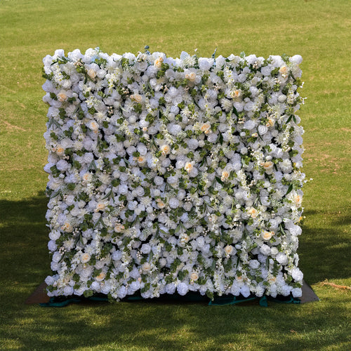 This is a beautiful artificial flower wall. It is densely covered with a variety of white flowers, including large, elegant roses and smaller blooms. Interspersed among the flowers are lush green leaves, creating a rich and textured appearance. The overall design is dense and full, giving a luxurious and abundant floral display, perfect for wedding or event backdrops.