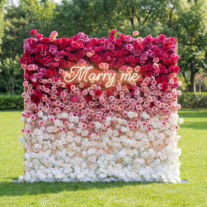 This is a romantic, gradient artificial flower wall backdrop from Valar Flowers, transitioning from deep red roses at the top to soft pink and pure white blooms at the bottom. A translucent "Marry me" sign overlays the center, set against a lush green lawn and trees, creating a dreamy, heartfelt setting perfect for marriage proposals, weddings, or special photo shoots.