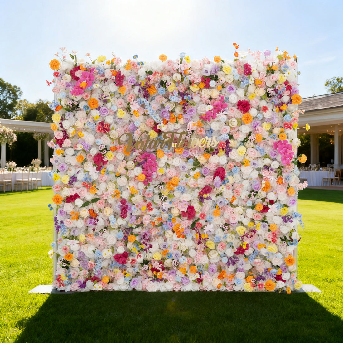 This image features a large, vibrant floral wall filled with an array of colorful flowers in shades of pink, yellow, orange, purple, and white. In the center, golden text reads &quot;Valoir Flowers&quot;. Set outdoors on a lush green lawn, with a bright blue sky and a elegant venue with tables and chairs in the background, it creates a lively and elegant atmosphere, perfect for events or decorative displays.