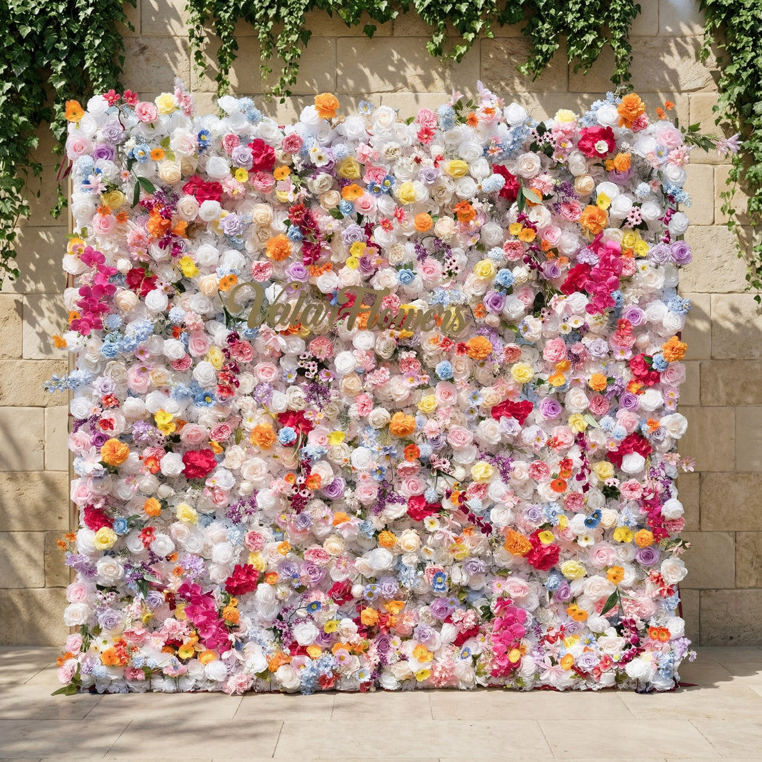 This is a stunning, dense artificial flower wall backdrop from Valar Flowers, bursting with a vibrant, rainbow-hued mix of lifelike blooms. Set against a stone wall with trailing ivy, it features creamy white roses, soft pink peonies, sunny yellow poppies, vivid orange carnations, sky-blue delphiniums, deep magenta orchids, and delicate filler flowers, creating a lush, romantic focal point perfect for weddings, photo booths, and luxury event decor.