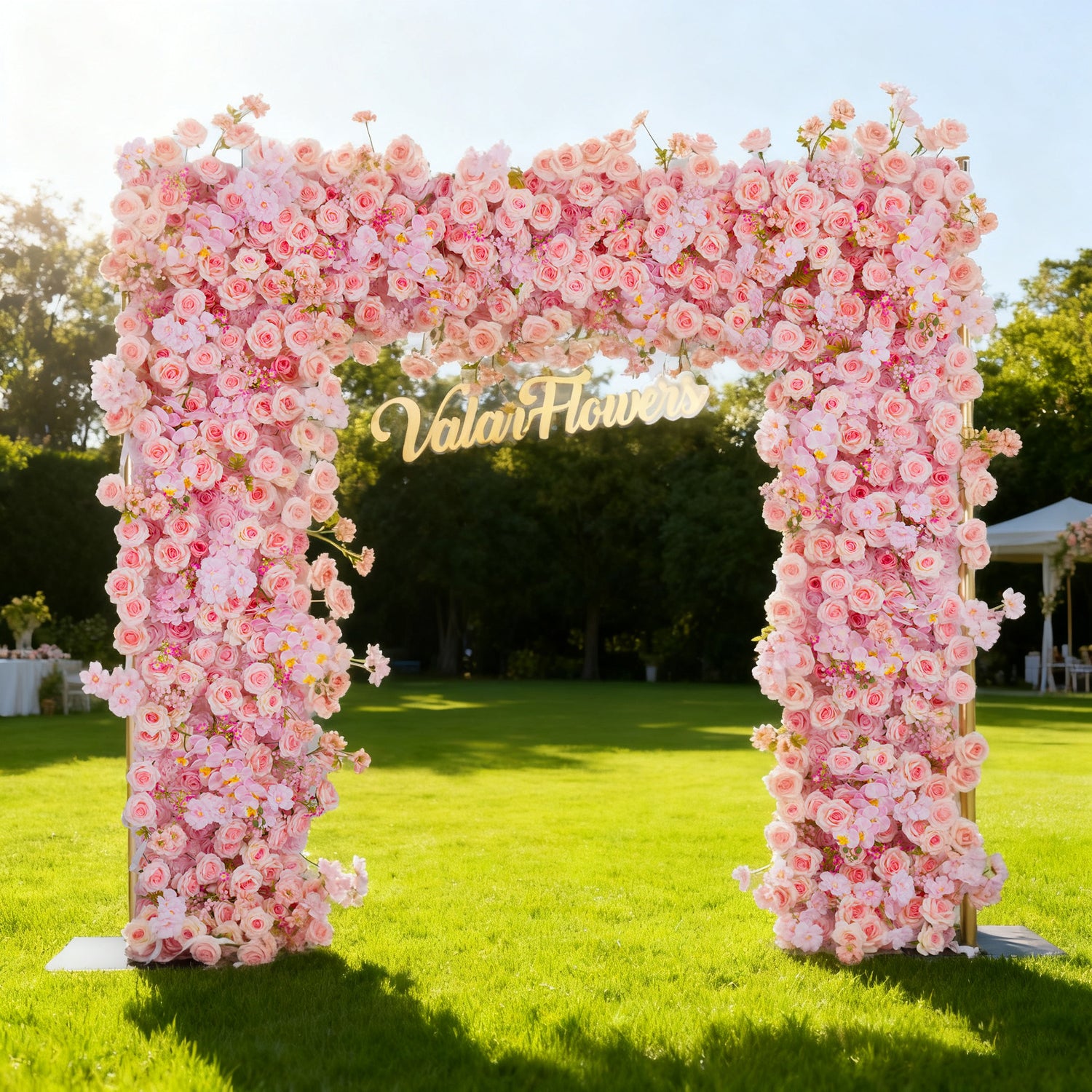 This image showcases a stunning floral arch by Valar Flowers, set outdoors on a lush lawn. It features a dense arrangement of artificial pink roses, with the brand’s name elegantly displayed in gold. Bathed in sunlight, with a scenic park and a white gazebo in the background, it exudes elegance, perfect for adding a luxurious, nature-inspired element to outdoor events like weddings.