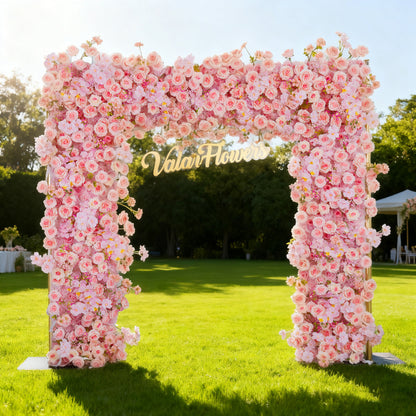 This image showcases a stunning floral arch by Valar Flowers, set outdoors on a lush lawn. It features a dense arrangement of artificial pink roses, with the brand’s name elegantly displayed in gold. Bathed in sunlight, with a scenic park and a white gazebo in the background, it exudes elegance, perfect for adding a luxurious, nature-inspired element to outdoor events like weddings.