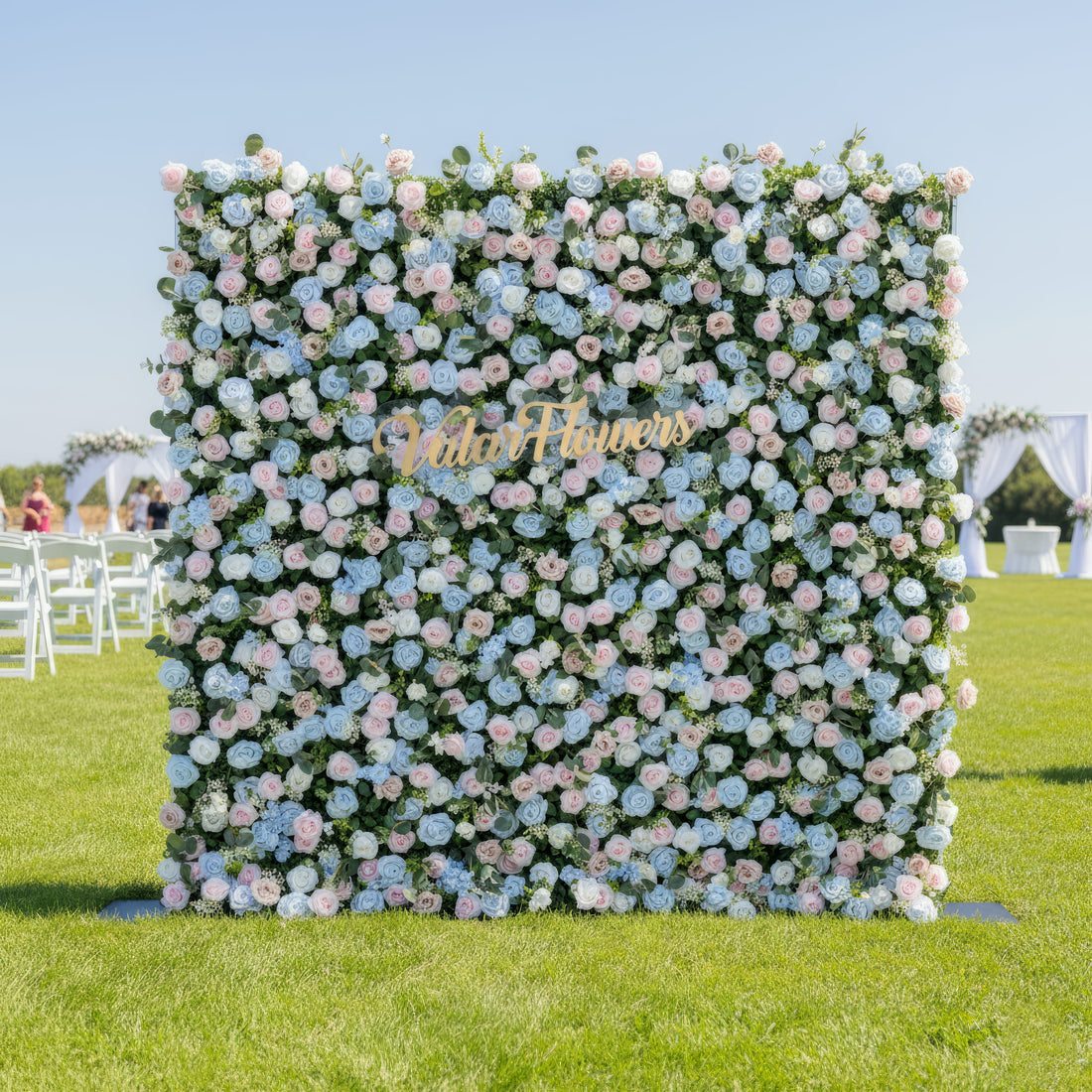 This image showcases a vibrant floral backdrop by Valar Flowers, set outdoors on a lush green lawn. It features a dense arrangement of artificial flowers in various shades of blue, light blue, and pink, with the brand’s name elegantly integrated in gold. In the background, there are event elements like white chairs and a floral arch, making it perfect for adding a sophisticated, nature-inspired touch to outdoor events such as weddings.