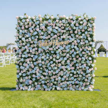 This image showcases a vibrant floral backdrop by Valar Flowers, set outdoors on a lush green lawn. It features a dense arrangement of artificial flowers in various shades of blue, light blue, and pink, with the brand’s name elegantly integrated in gold. In the background, there are event elements like white chairs and a floral arch, making it perfect for adding a sophisticated, nature-inspired touch to outdoor events such as weddings.