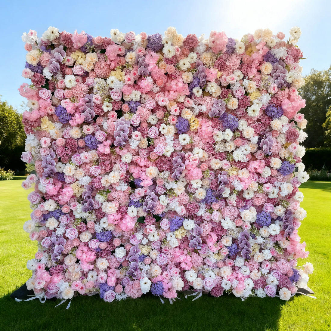 This image showcases a large, vibrant floral wall brimming with an array of colorful flowers in shades of pink, purple, white, and yellow. Set outdoors on a lush green lawn under a bright sky, it creates a lively and cheerful atmosphere, perfect for decorative displays or events, exuding a sense of natural beauty and joy.