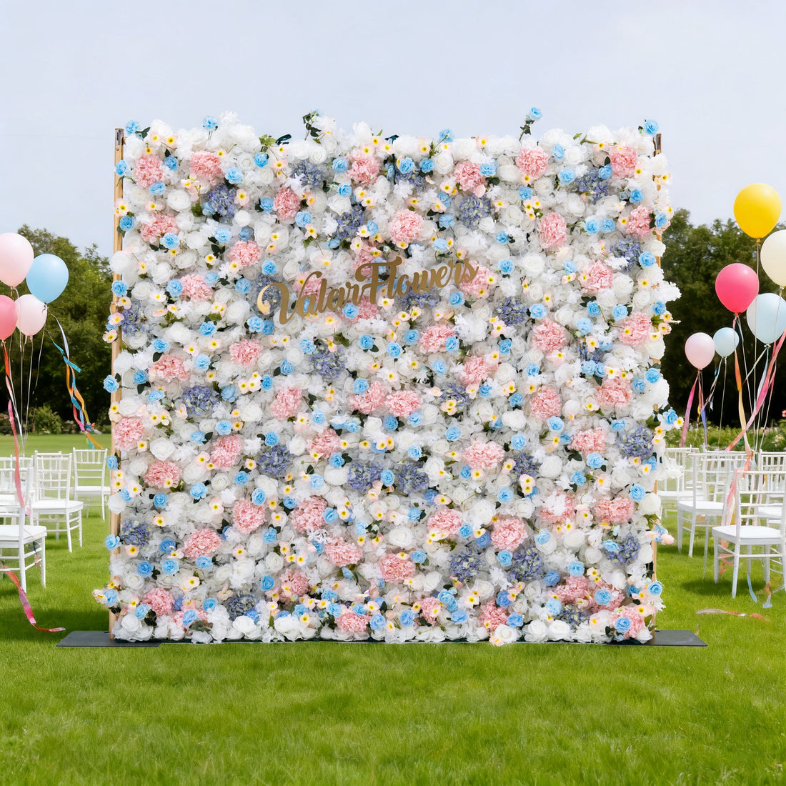 This image features a large floral wall with a soft palette of white, pink, and blue flowers. &quot;ValarFlowers&quot; is displayed in golden text on it. Set outdoors on a lush green lawn, the scene includes white chairs and colorful balloons, creating a cheerful and elegant atmosphere, perfect for events like weddings or celebrations.
