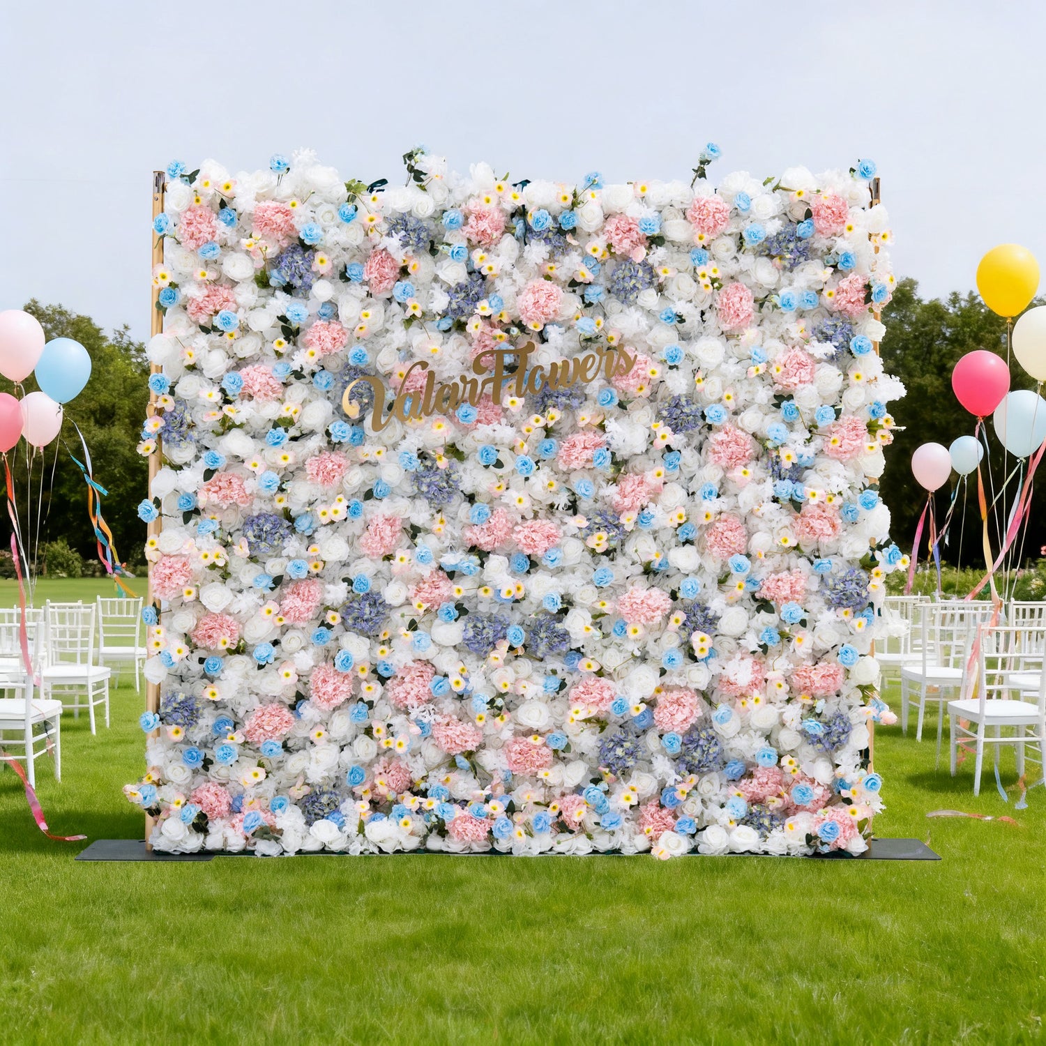 This image features a large floral wall with a soft palette of white, pink, and blue flowers. &quot;ValarFlowers&quot; is displayed in golden text on it. Set outdoors on a lush green lawn, the scene includes white chairs and colorful balloons, creating a cheerful and elegant atmosphere, perfect for events like weddings or celebrations.