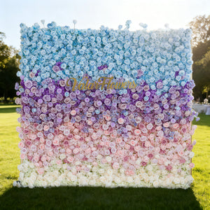 This image features a large floral wall with a gradient of colors from blue at the top, transitioning through purple, pink, to white at the bottom. "Valar Flowers" is displayed in golden text. Set outdoors on a lush green lawn under a bright sky, it creates a vibrant and elegant atmosphere, perfect for events or decorative displays.