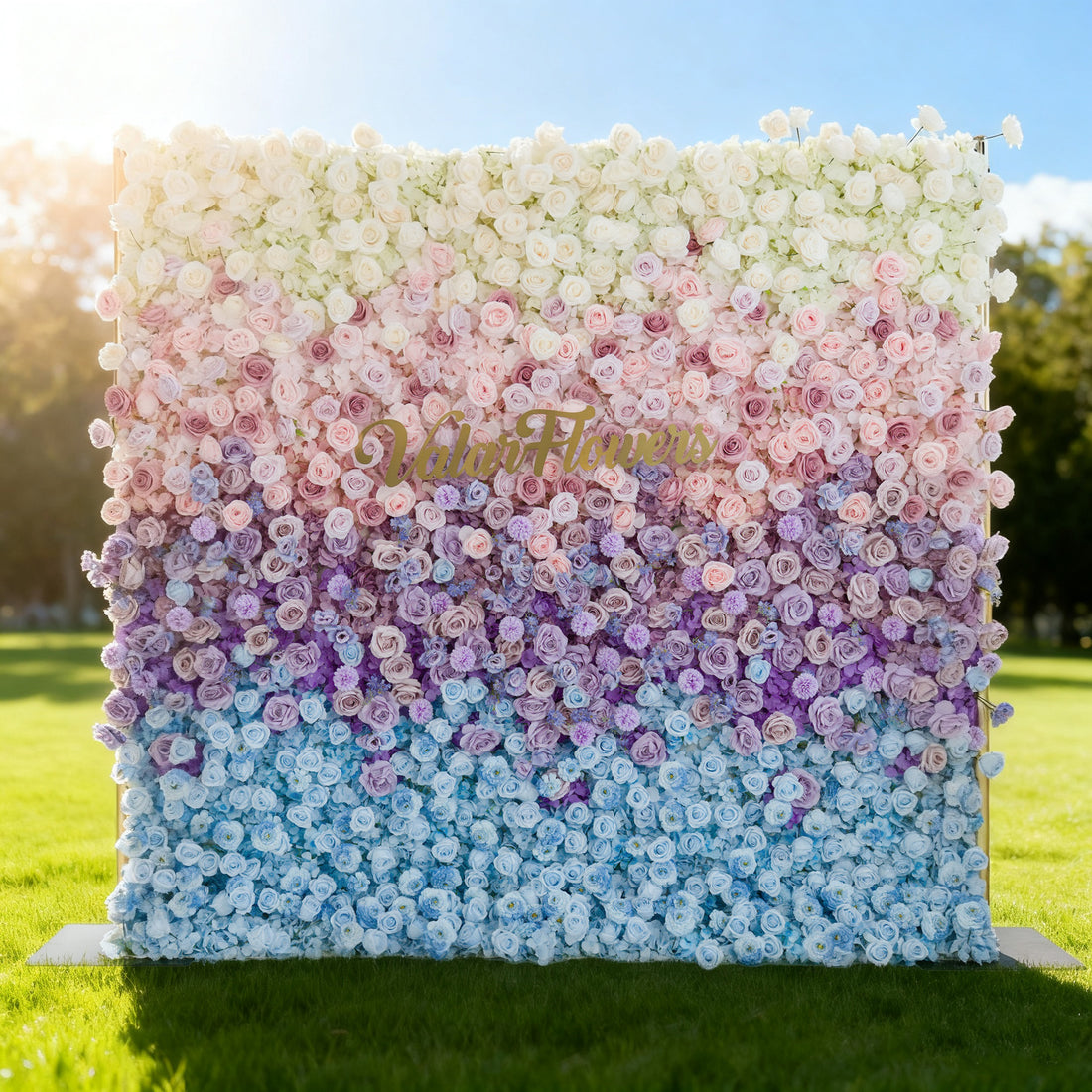 This image showcases a large floral wall with a beautiful gradient of colors, transitioning from white and pink at the top to purple and blue at the bottom. &quot;Valar Flowers&quot; is displayed in golden text. Set outdoors on a lush green lawn under a bright sky, it creates a vibrant and elegant atmosphere, perfect for events or decorative displays.