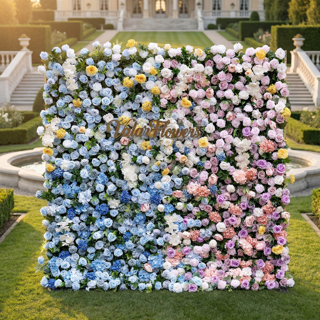 This is ValarFlowers’ premium gradient artificial flower wall backdrop, set against a luxurious European-style garden with a grand staircase and fountain. It features a seamless color transition from sky-blue delphiniums at the base to creamy white roses, sunny yellow poppies, blush pink peonies, and lavender roses at the top. This lifelike, reusable floral arrangement creates an elegant, romantic focal point ideal for weddings, photo booths, bridal showers, and luxury event decor.