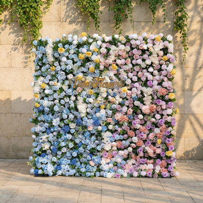 This is a stunning, gradient-style artificial flower wall backdrop from Valar Flowers, set against a sunlit stone wall with trailing ivy. It features a lush, dense arrangement of lifelike blooms: soft sky-blue delphiniums at the bottom, transitioning upward to creamy white roses, blush pink peonies, sunny yellow poppies, and lavender roses. The seamless color gradient creates an elegant, romantic focal point perfect for weddings, photo booths, and luxury event decor.
