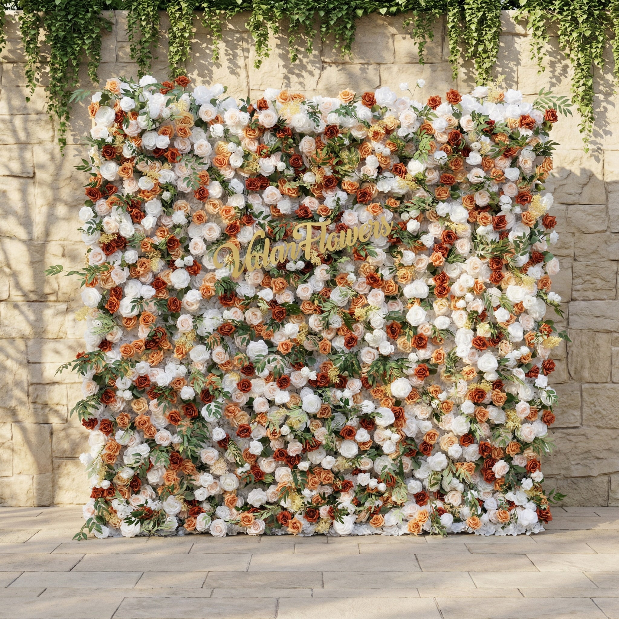 This is ValarFlowers’ premium terracotta &amp; cream artificial flower wall backdrop, set against a sunlit stone wall with trailing ivy. It features a lush, dense arrangement of lifelike blooms: burnt orange roses, creamy white peonies, blush pink roses, and vibrant green foliage. This warm, timeless, reusable floral display creates an elegant, romantic focal point ideal for rustic weddings, fall events, photo booths, and luxury outdoor decor.