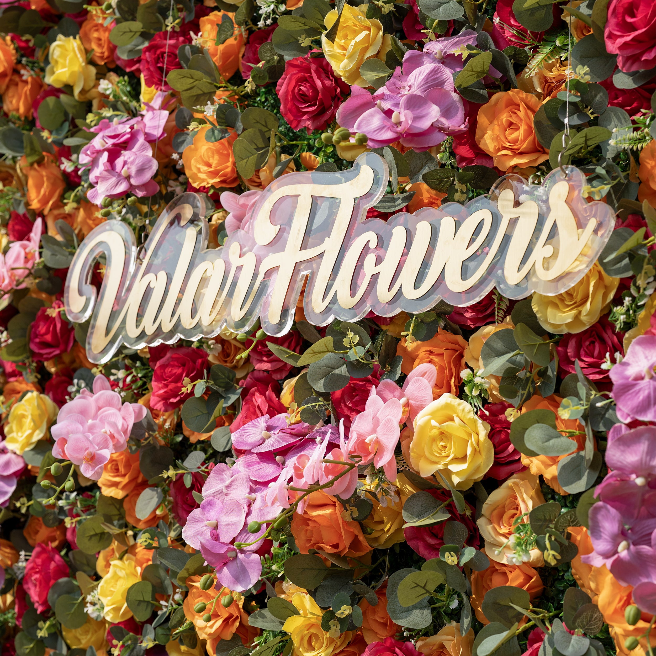 This is a close-up of ValarFlowers’ premium multicolor artificial flower wall, featuring a lush, dense arrangement of lifelike red, orange, yellow roses, pink phalaenopsis orchids, and realistic eucalyptus greenery. Centered is a custom acrylic "ValarFlowers" logo sign. This high-quality, reusable floral backdrop delivers a vibrant, fresh aesthetic, ideal for wedding backdrops, photo booths, summer events, and luxury event decor.