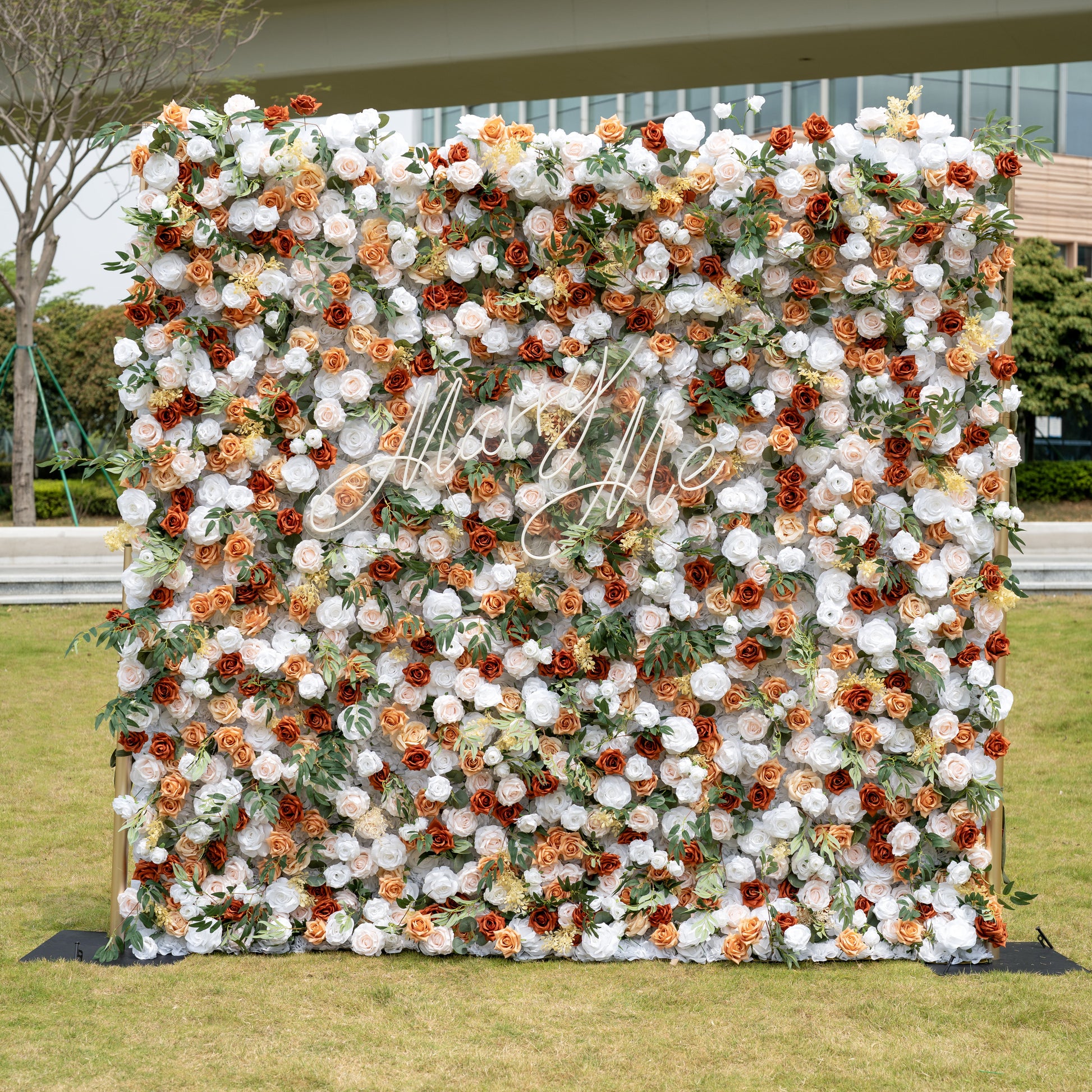 This is a stunning 8x8ft (2.4x2.4m) artificial flower wall backdrop, mounted on a golden metal stand and set outdoors on a grassy lawn with an urban skyline in the background. It features a lush, dense arrangement of lifelike blooms in a warm terracotta and cream palette: burnt orange roses, creamy white peonies, soft peach roses, and vibrant green foliage, centered with an elegant "Marry Me" neon script sign. This premium, reusable floral display creates a romantic, showstopping focal point, perfect for we