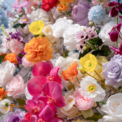This is a lush, vibrant close-up of a premium artificial mixed-flower arrangement, brimming with lifelike blooms in a joyful, colorful palette. It features creamy white roses, soft pink peonies, sunny yellow poppies, vivid orange carnations, sky-blue pom-pom flowers, deep pink orchids, lavender roses, and delicate purple filler flowers. The layered, realistic petals create a dreamy, romantic, whimsical aesthetic, perfect for wedding backdrops, event decor, and photo booth styling.