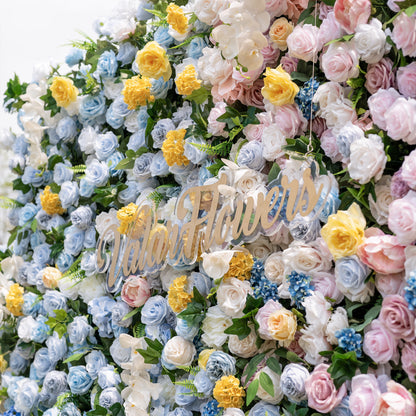 This is a stunning, ultra-dense artificial flower wall from ValarFlowers, bursting with lifelike blooms in a dreamy pastel gradient. It features sky-blue roses, creamy white roses, sunny yellow carnations, blush pink peonies, lavender roses, and lush green foliage, with the brand’s elegant gold script logo at the center. This premium, reusable floral arrangement creates a romantic, showstopping focal point ideal for weddings, photo booths, bridal showers, and luxury event decor.