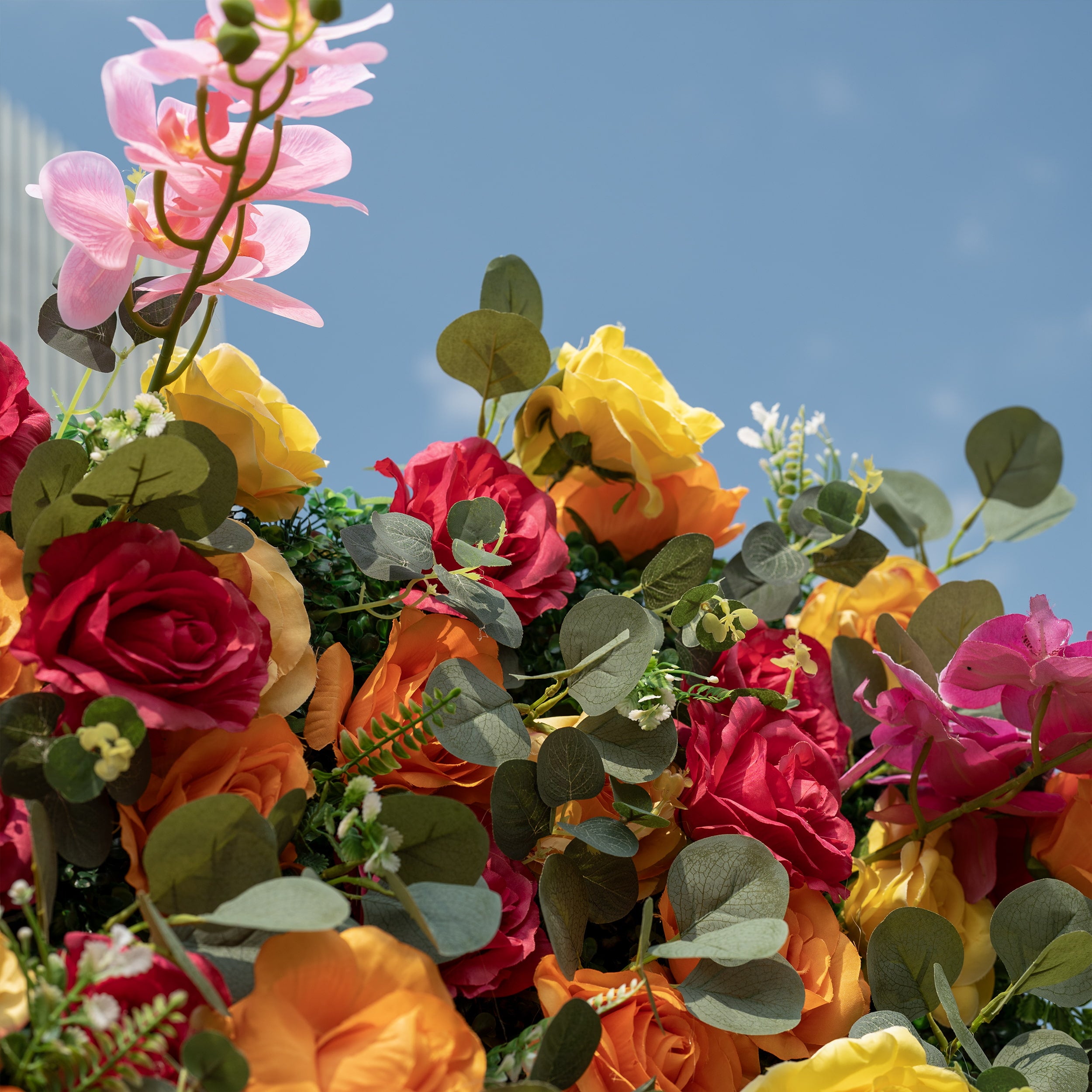 This is a close-up of a premium artificial flower wall arrangement, brimming with lifelike blooms in a vibrant multicolor palette. It features red, orange, yellow roses, delicate pink phalaenopsis orchids, and realistic eucalyptus greenery. High-quality, realistic petals deliver a fresh, lush aesthetic, perfect for wedding backdrops, photo booths, summer events, and luxury event decor.