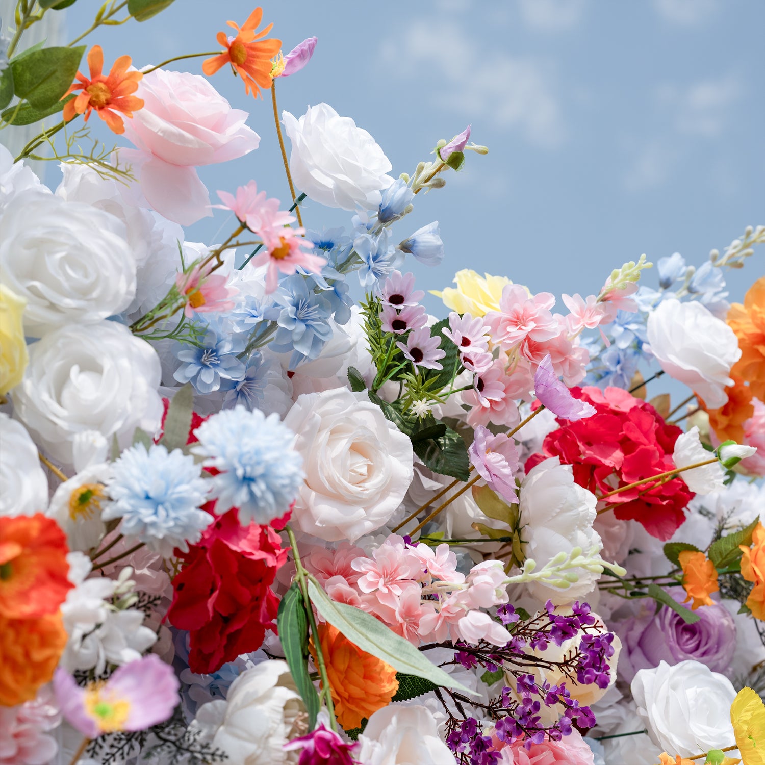 Valar Flowers premium vibrant mixed artificial flower wall backdrop, a lush, layered arrangement of lifelike blooms including white roses, pink peonies, orange carnations, blue delphiniums, red hydrangeas, and delicate filler flowers. Set against a bright blue sky, this dense, colorful floral display creates a dreamy, romantic focal point perfect for outdoor weddings, engagement parties, photo booths, and luxury event decor.