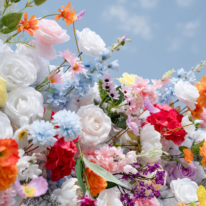 Valar Flowers premium vibrant mixed artificial flower wall backdrop, a lush, layered arrangement of lifelike blooms including white roses, pink peonies, orange carnations, blue delphiniums, red hydrangeas, and delicate filler flowers. Set against a bright blue sky, this dense, colorful floral display creates a dreamy, romantic focal point perfect for outdoor weddings, engagement parties, photo booths, and luxury event decor.