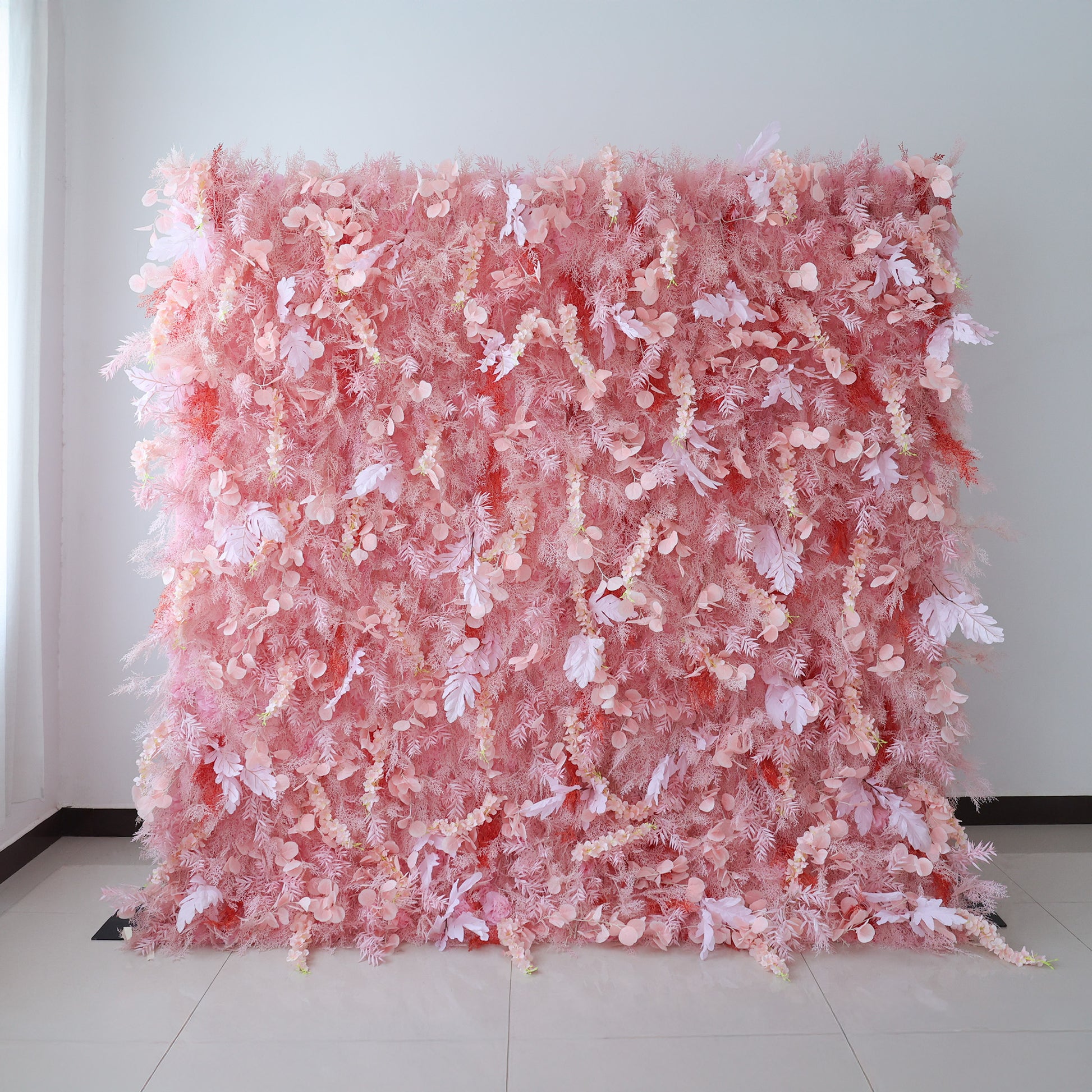 This image showcases a large, dense floral wall indoors. It's covered with an array of delicate pink flowers and foliage, creating a soft and romantic display. Set against a plain wall with a tiled floor, the harmonious blend of various pink hues and intricate floral elements exudes a sense of elegance and gentle charm, perfect for decorative purposes in events or as a striking interior backdrop.
