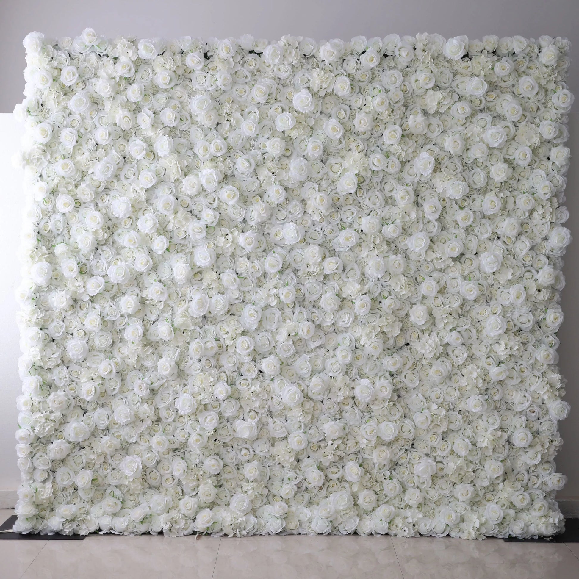 This opulent indoor floral backdrop features a dense, wall-to-wall arrangement of artificial white roses and delicate filler blooms, creating a lush, velvety texture. Set against a plain light-colored tiled floor, its pure, monochromatic design exudes timeless elegance and romance, making it a sophisticated focal point for weddings, engagement photos, or upscale events.