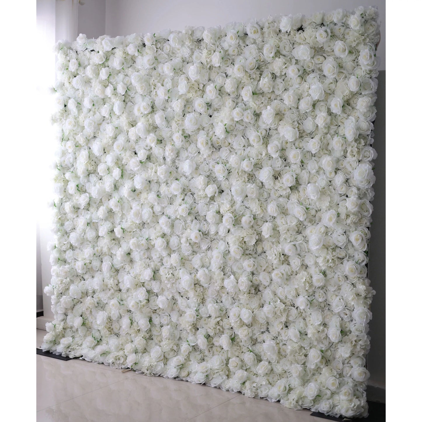 This striking indoor floral backdrop features a dense, wall-to-wall arrangement of artificial white roses and delicate filler blooms, creating a lush, velvety texture. Set against a plain white wall and light-colored tiled floor, its pure, monochromatic design exudes timeless elegance and romance, making it a sophisticated focal point for weddings, engagement photos, or upscale events.