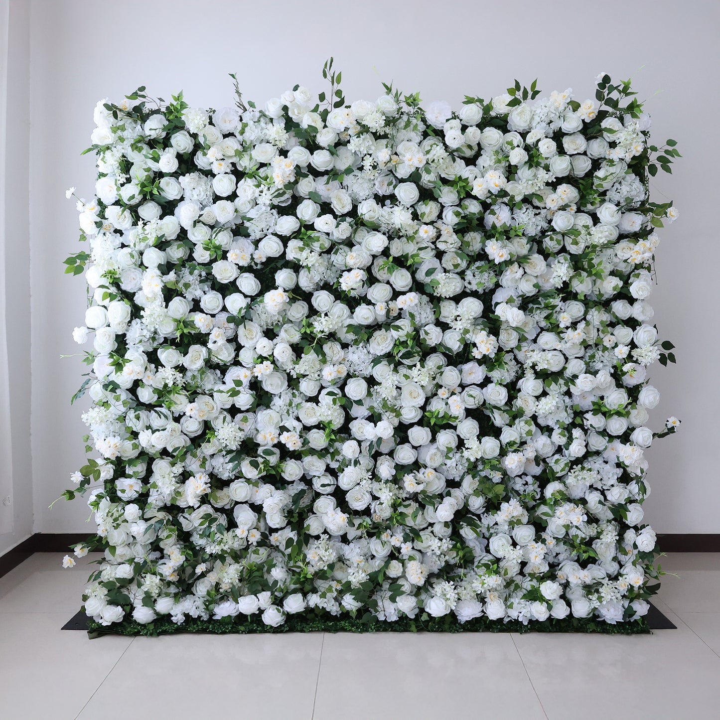 This image showcases a large, square floral wall indoors. It's densely covered with an array of white flowers, creating a pure and elegant display. Set against a plain wall with a tiled floor, the scene exudes a sense of purity and charm, perfect for decorative purposes in events or as a striking interior backdrop.