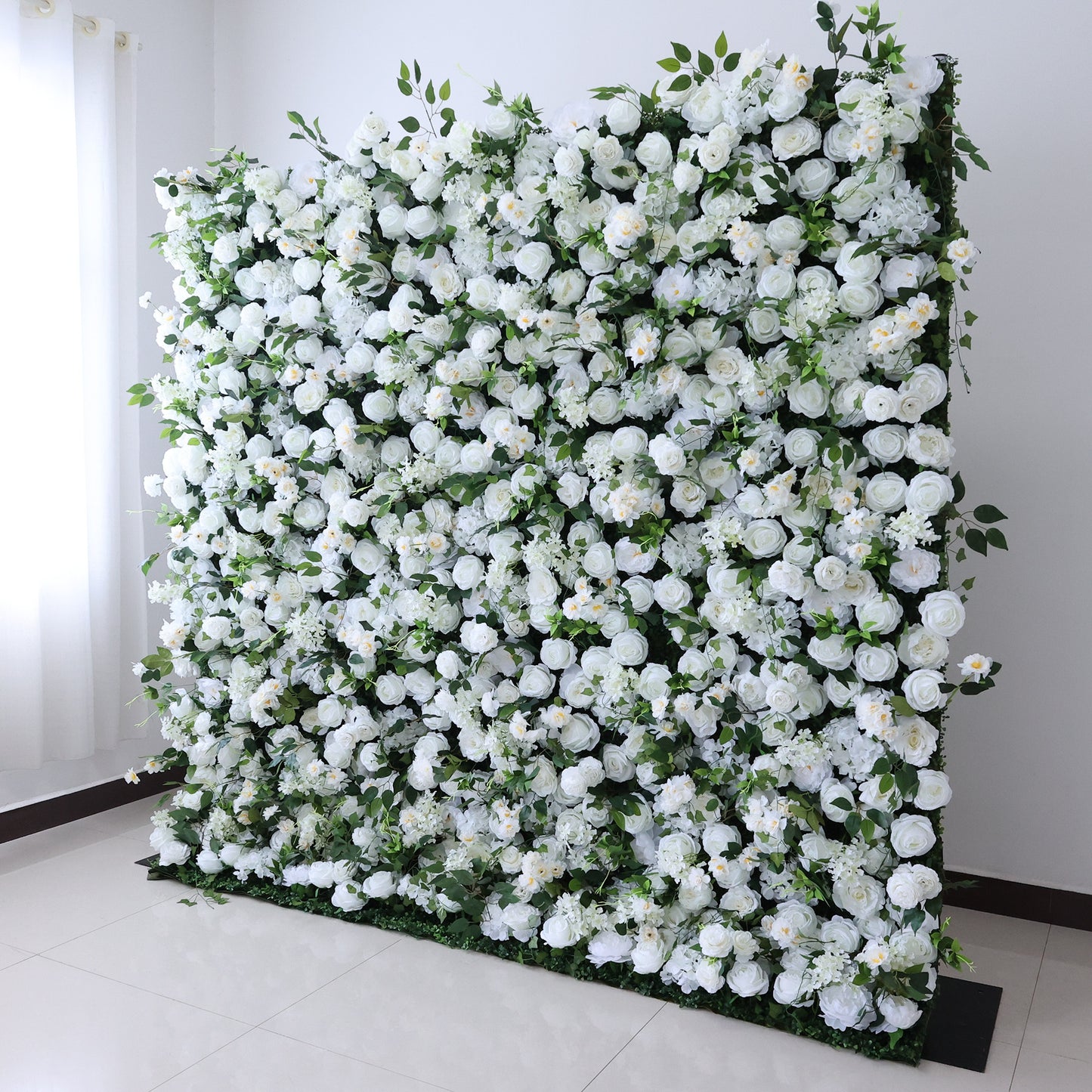This image showcases a large, artificial floral wall indoors. It's densely covered with an array of white flowers, creating a pure and elegant display. Set in a room with white walls and a tiled floor, the scene exudes a sense of purity and charm, perfect for decorative purposes in events or as a striking interior backdrop.