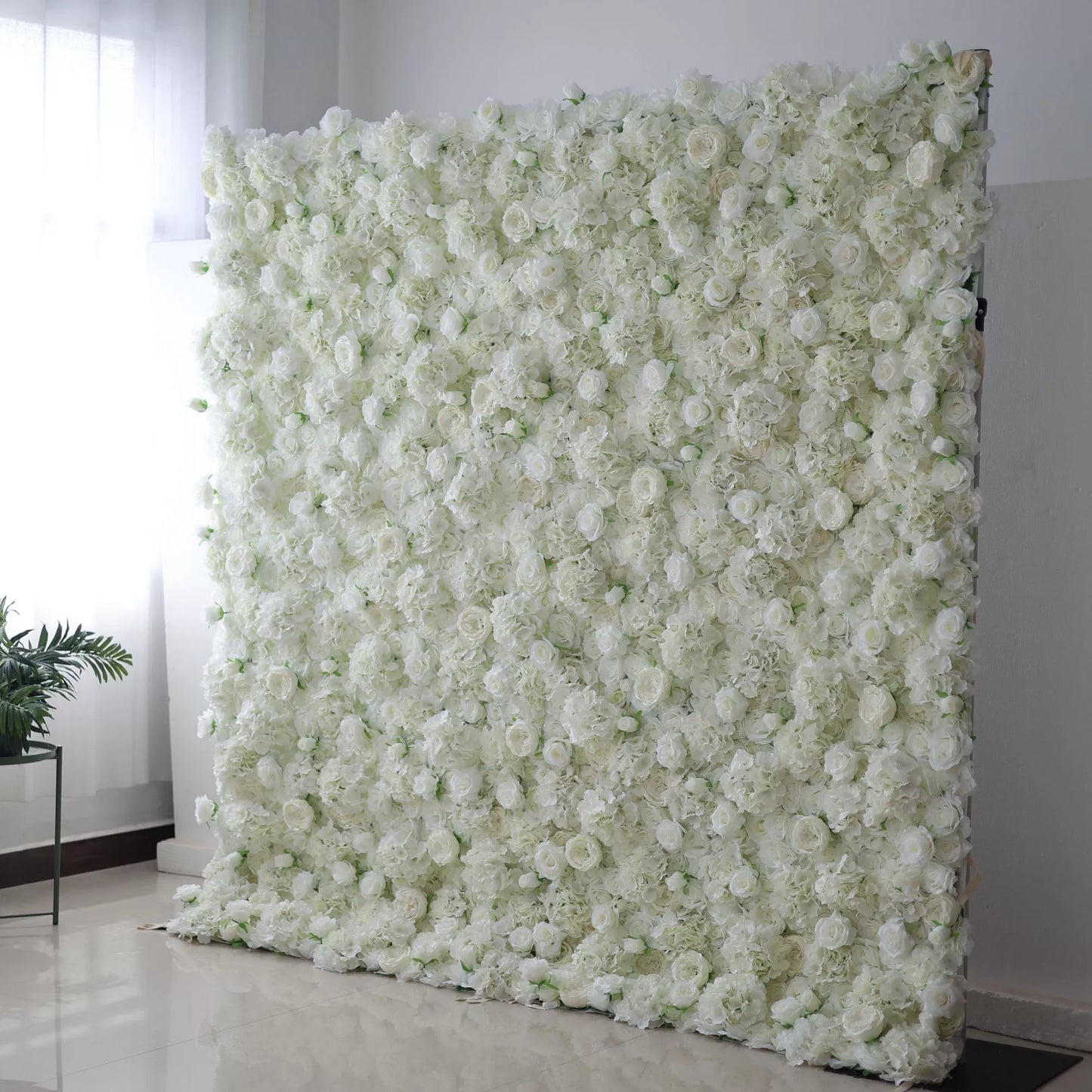 This image features a large, square floral wall indoors. It's densely covered with pure white flowers, creating a clean, elegant, and serene display. The flowers, including roses and other blooms, are tightly packed, giving a sense of opulence and purity. Set against a plain white wall with a shiny floor, and with a potted plant nearby, the scene exudes an atmosphere of sophistication and natural beauty, perfect for decorative purposes in events or as a striking interior backdrop.