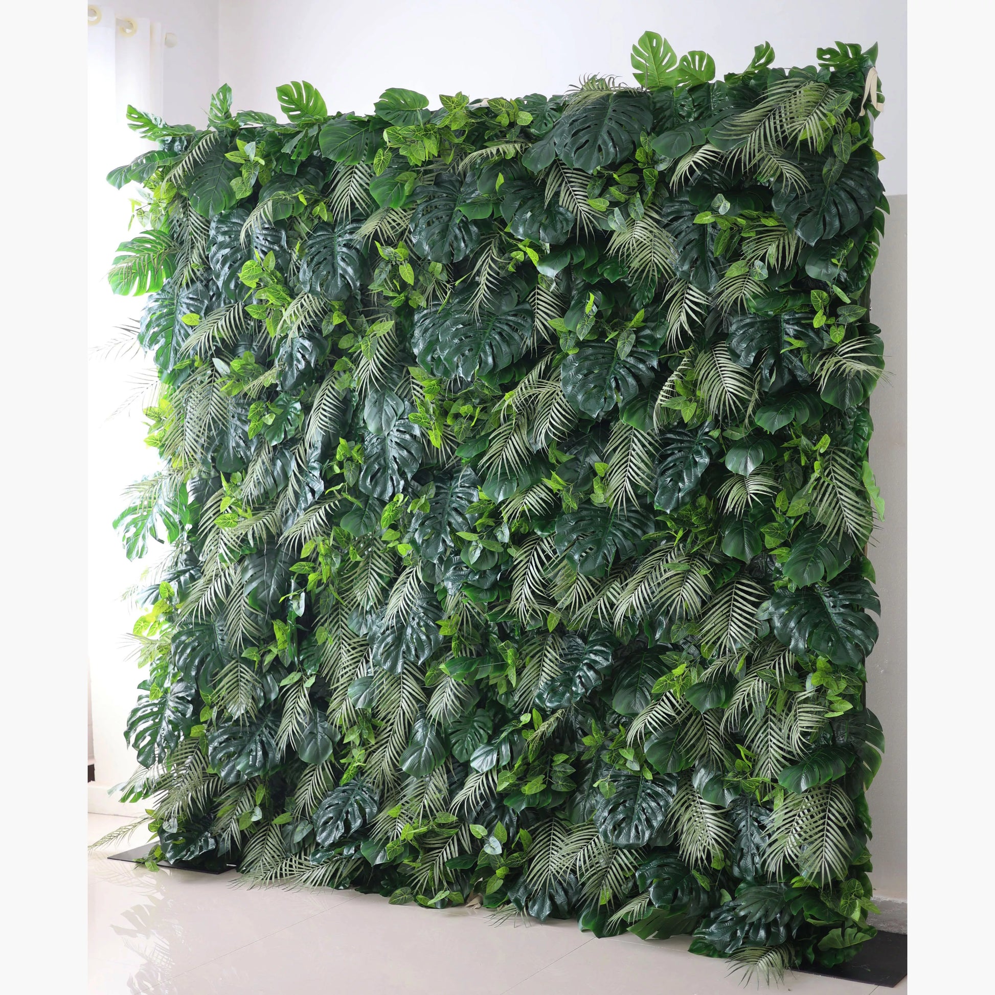 Valar Flowers' Artificial Fabric Green Wall: Tropical Eden. A large artificial greenery wall showcases a dense mix of lush leaves. Big monstera leaves and delicate fern - like fronds in various shades of green create a vibrant, tropical - inspired texture. It’s a fresh, natural - looking backdrop, perfect for adding an organic touch to decor or events. 