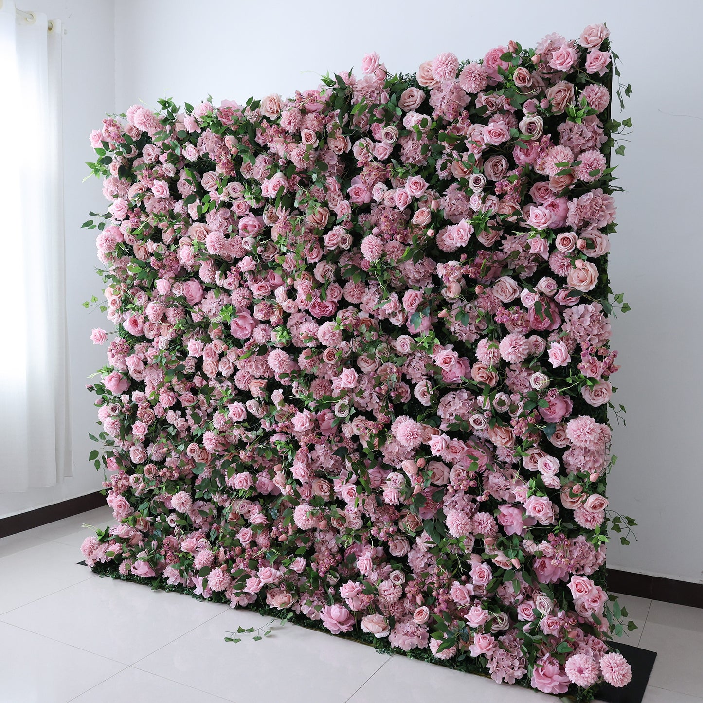 This image features a large, artificial floral wall indoors. It's densely covered with an array of pink flowers, creating a rich, romantic display. Set in a room with white walls and a tiled floor, the scene exudes a sense of elegance and charm, perfect for decorative purposes in events or as a striking interior backdrop.
